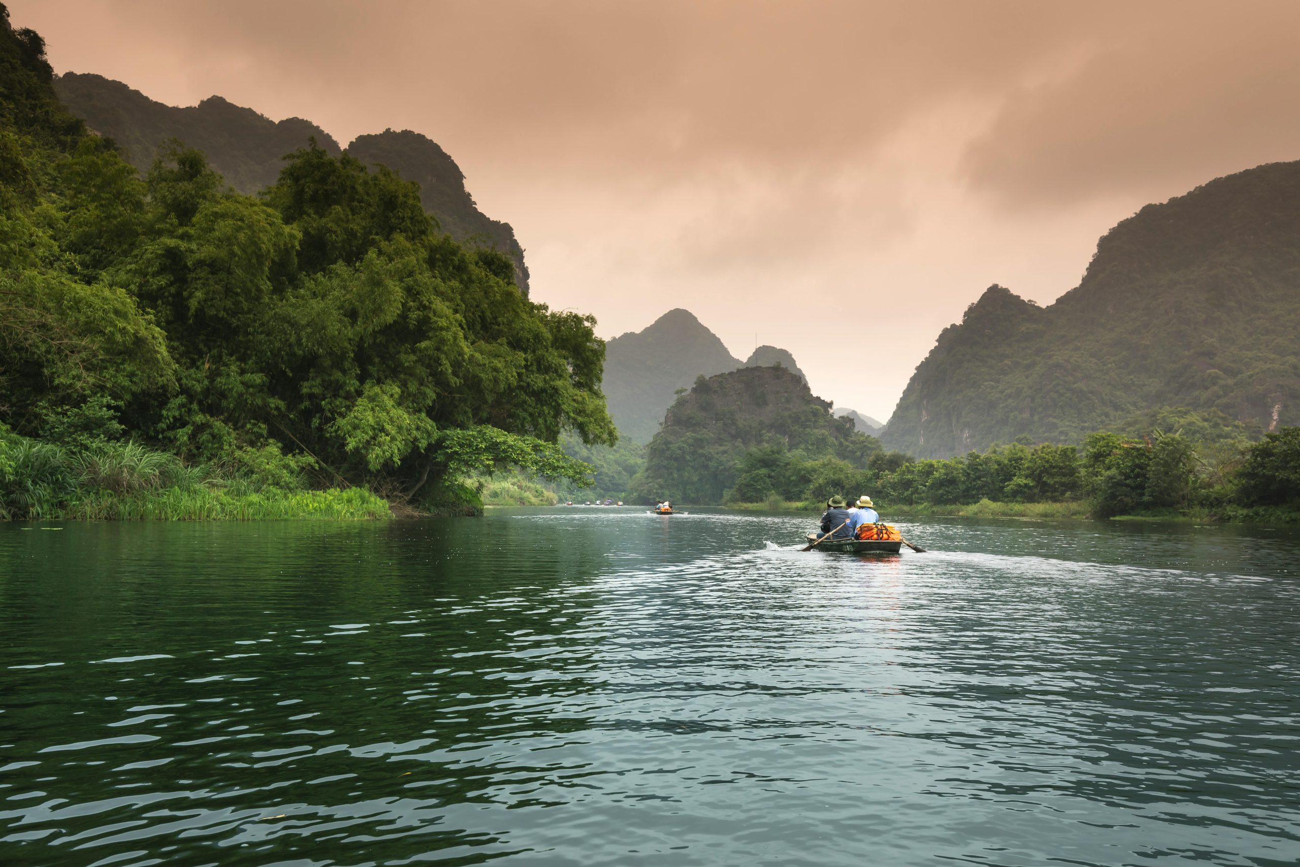 Two people rowing a boat through a serene river surrounded by lush greenery and mountains.