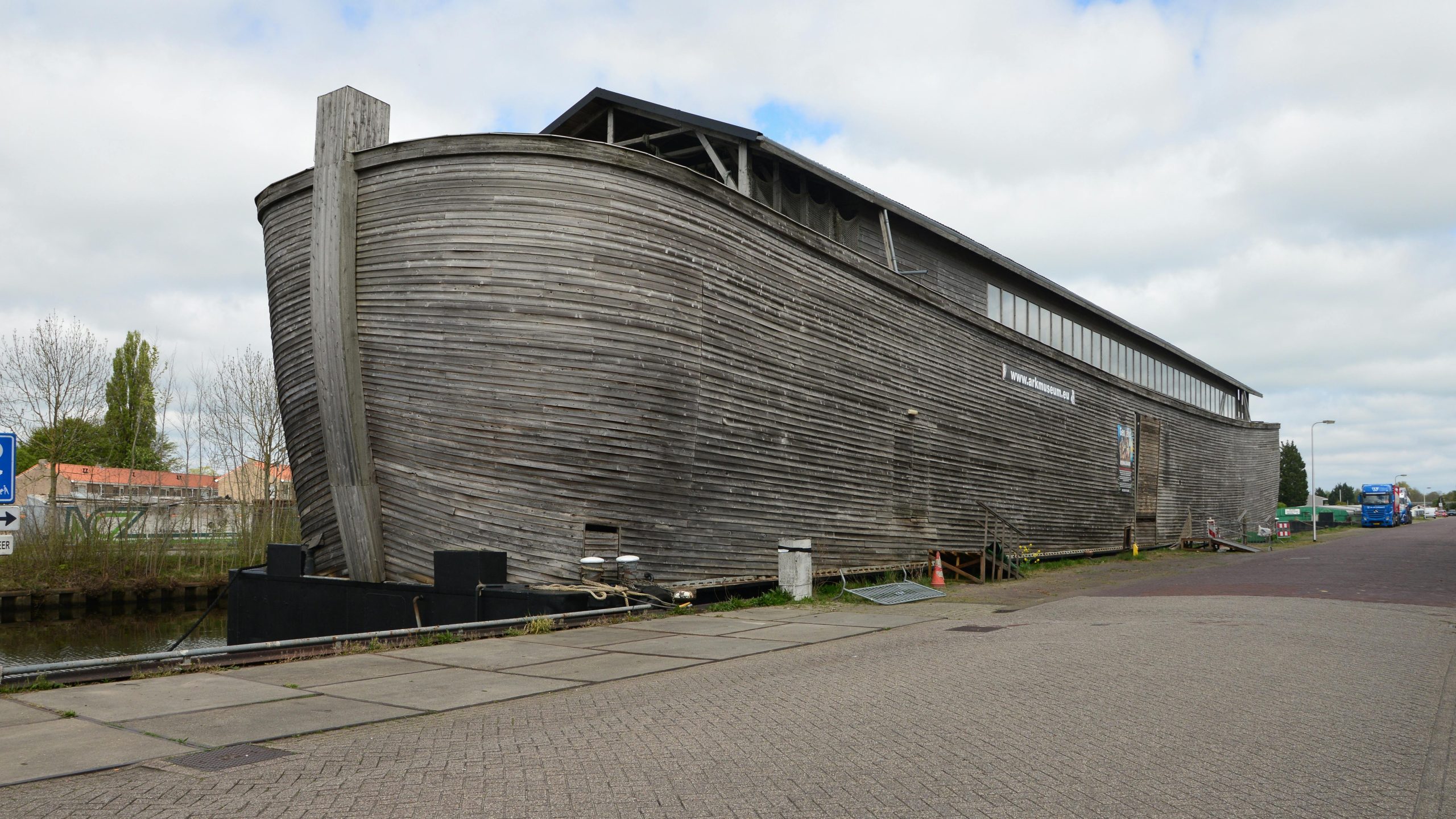 Large wooden structure by canal, resembling a historical ark on a cloudy day.