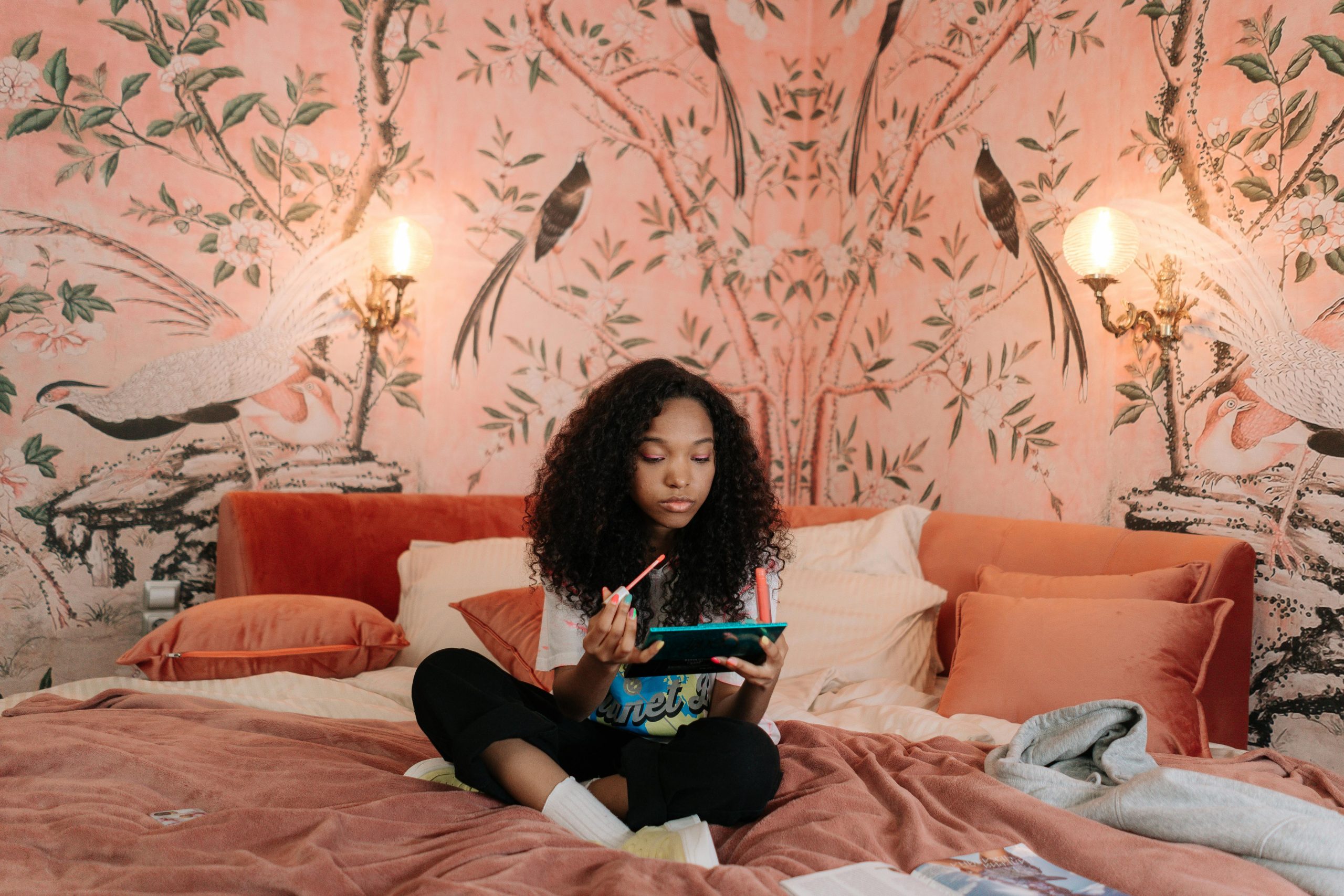 Young girl with curly hair applies makeup while sitting on a bed in a chic, artful bedroom. Cozy and fashionable setting.