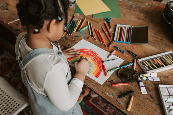 Young child creating a vibrant rainbow drawing at a wooden table with various art supplies.